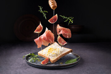 Crostini with prosciutto ricotta and figs, flying Italian ham and fruit on a dark background, toast...