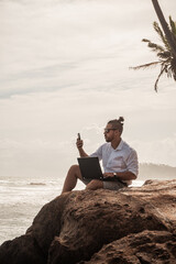 Cyfrowy nomada, człowiek pracujący z laptopem i smartfonem zdalnie na tle oceanu i skał...