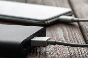 The smartphone is charging from external powerbank with cable on a brown wooden background. Closeup, selective focus