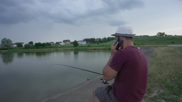adult man fisherman in hat sits with fishing rod on chair on shore of reservoir and talks by phone in summer cloudy day