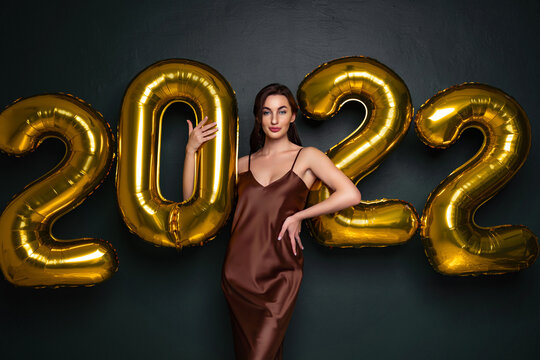 Happy young brunette woman in glamour dress near golden 2022 balloons on black background. New Year celebration. Looking in the camera. standing between the numbers air balloons