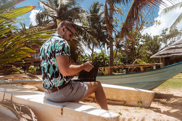 Fototapeta na wymiar Cyfrowy nomada, mężczyzna pracujący na tropikalnej plaży z laptopem i telefonem.