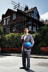 Man wearing suit, holding hard hat, standing outside house with scaffolding