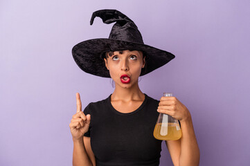 Young mixed race woman disguised as a witch holding potion isolated on purple background  pointing upside with opened mouth.