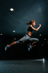 Fototapeta na wymiar Slender woman is actively jumping high in sports clothes on a black background of the gym. Dynamic movement side view.