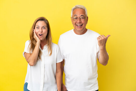 Middle age couple isolated on yellow background pointing finger to the side and presenting a product