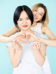 Love sign. Happy women. Ethnic diversity. Tolerance acceptance. Best friends. Pretty smiling asian caucasian ladies showing heart shaped gesture isolated blue.