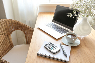 Home office desk with laptop computer, calculator, spiral book, coffee and a white flower bouquet,...