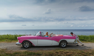 Young man pushing vintage convertible at coast whilst girlfriend drives, Havana, Cuba - obrazy, fototapety, plakaty