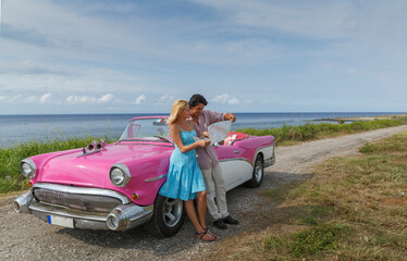 Young couple looking at map beside vintage convertible at coast, Havana, Cuba - obrazy, fototapety, plakaty