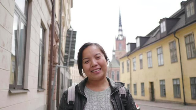 Front view of happy Asian woman standing and smiling on the street in town, going out for a walk on the street in Sweden. Traveling abroad on long holiday. Looking at camera concept