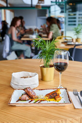 View of a plate of pork tenderloin with grilled pineapple accompanied by a glass of red wine at a restaurant table with people in the background out of focus.