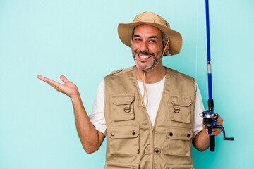 Middle age caucasian fisherman holding rod isolated on blue background  showing a copy space on a palm and holding another hand on waist.