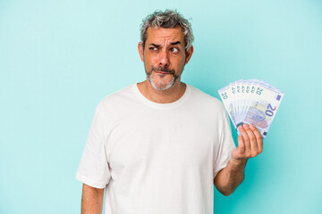 Middle age caucasian man holding bills isolated on blue background  confused, feels doubtful and unsure. - obrazy, fototapety, plakaty
