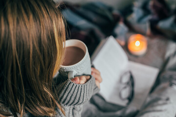 Young girl drinking winter beverage hot cacao, sitting on cozy bed, reading book. Cozy warm image