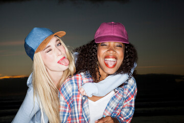 Young woman wearing baseball cap arm around friend looking at camera sticking out tongue