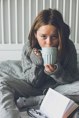 Young girl drinking hot chocolate on cozy bed in bedroom. Concept of autumn or winter weekend