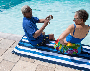 Senior couple sitting beside swimming pool, man taking photograph of woman using smartphone