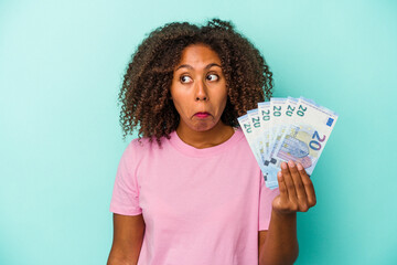Young african american woman holding euro banknotes isolated on blue background shrugs shoulders...