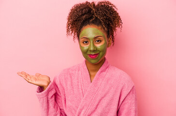 Young african american woman wearing a bathrobe and facial mask isolated on pink background showing a copy space on a palm and holding another hand on waist.