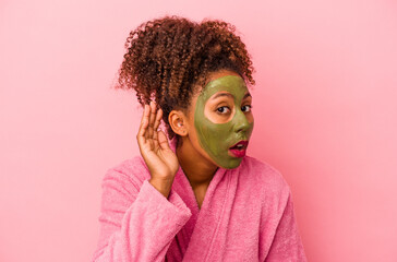 Young african american woman wearing a bathrobe and facial mask isolated on pink background trying to listening a gossip.