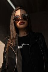 Stylish portrait of a beautiful young woman model in fashionable black leather clothes with a jacket and vintage sunglasses outdoors. Creative photo with sun light and shadow