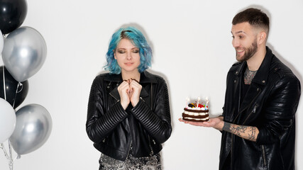 Birthday wish. Happy beloved couple. Festive surprise. Excited man presenting holiday cake with candle lights dreamful woman isolated white black silver balloons decor.