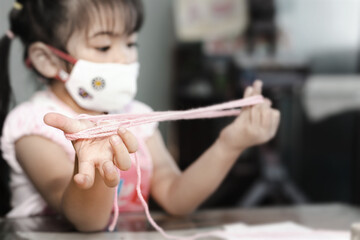 Little Asian girl wearing facial hygienic mask playing cats cradle game at home,selective focus....