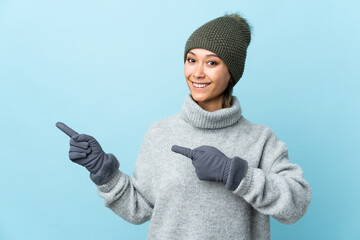 Young Uruguayan girl with winter hat isolated on blue background pointing finger to the side