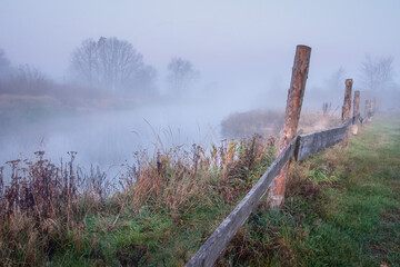 Obraz premium Scenery of autumn nature on misty sunrise. Road near misty river. Dry grass along the river bank.