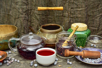 Japanese vintage rustic copper tea kettle and clear glass Japanese teapot, tea cup filled with red hibiscus tea, other items and juniper berries scattered about on wood surface