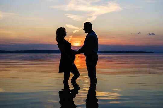 Silhouette of a married couple stands in the water of the lake against the background of sunset. The family is expecting a baby.