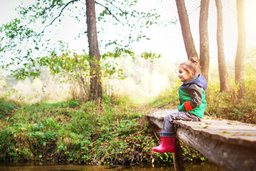 Naklejka na ściany i meble A child in red rubber boots sits on a wooden bridge over the river and looks at the water. Autumn landscape, cold weather, dry fallen leaves. Autumn mood, carefree childhood
