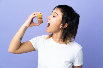 Young Uruguayan woman over isolated purple background holding colorful French macarons and eating it