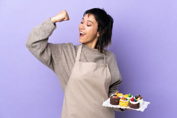 Young Uruguayan woman holding lots of different mini cakes over isolated purple background celebrating a victory