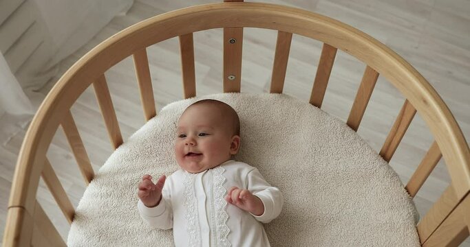 Top view cute four-month-old newborn baby in white bodysuit lying down in comfy mattress of cosy wooden crib. Store ad of comfortable modern furniture for infants, healthy babyhood, childcare concept