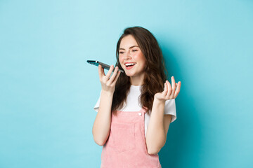Portrait of smiling woman holding phone near lips and talking, using app translator on smartphone or recording voice message, speaking with speakerphone, standing over blue background - obrazy, fototapety, plakaty