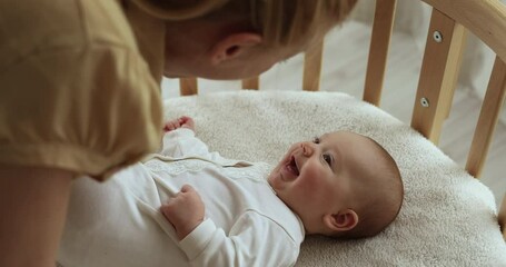 Loving mother amuse to baby lying on comfort crib, close up. Cheery lively adorable infant looks at caring mommy, playing having fun spend priceless time together at home. Care and motherhood concept