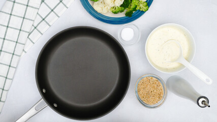Cauliflower and broccoli casserole recipe. Frying pan, vegetables, Alfredo sauce, and bread crumbs close up on kitchen table, flat lay