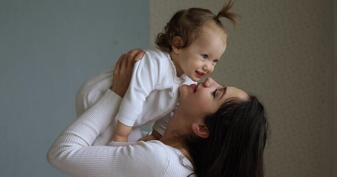 Loving young mother lifts on arms raises and kisses her cute infant baby girl, enjoy playtime having fun together at home. Concept of childcare and cherish, happy life moments with child, affectionate