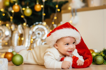 Cute smiling baby is lying under a festive Christmas tree and playing with gifts. Christmas and New Year celebrations