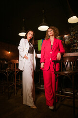 Two beautiful women of Caucasian ethnicity in bright stylish suits and evening makeup, in the evening at the bar, fashionably posing for the camera