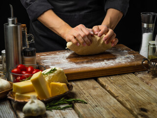 The chef kneads the dough on a cutting board. Cooking Italian focaccia bread, pizza. Ingredients - cheese, garlic, tomatoes, basil. Wooden texture. Restaurant, hotel, bakery, home cooking.