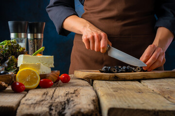 The process of preparing vitamin salad Greek. Lots of fresh bright ingredients on the table. The chef cuts olives on a cutting board. Beautiful composition. Macro photography.