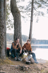 A married couple prepares food on a campfire on the shore of the lake with a tent.