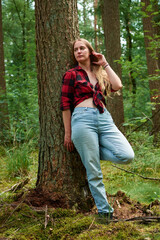 Naklejka na ściany i meble A young Italian woman wearing a red flannel with jeans and leaning on a tree in a forest