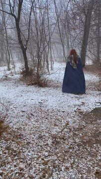 Magical woman with red dreadlocks in viking costume with hatchet and mantle goes into snow forest. Amateur shooting of cosplayer's retreating person from back. Winter atmosphere in wood with river.