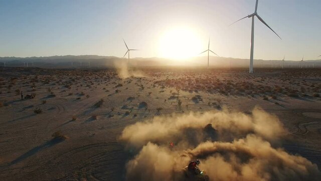 4k Aerial of ATV riding through dust cloud donuts with wind turbine sunset