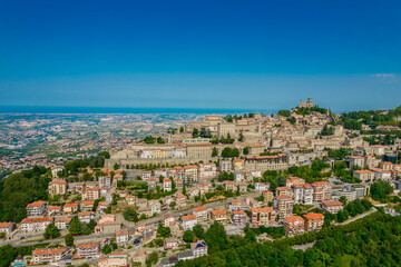 Fototapeta na wymiar Aerial view of San Marino old town with old buildings and red roofs on the hill on a sunny day with clear sky. Picture from above with Italy behind in horizon