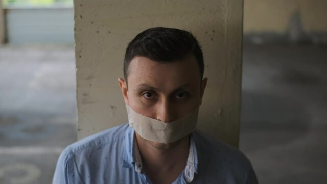businessman with his mouth sealed with tape sits tied to post, looks up at camera. Seen man in blue shirt, victim of maniac with gag in his mouth, cannot speak, dumb. Rack focus